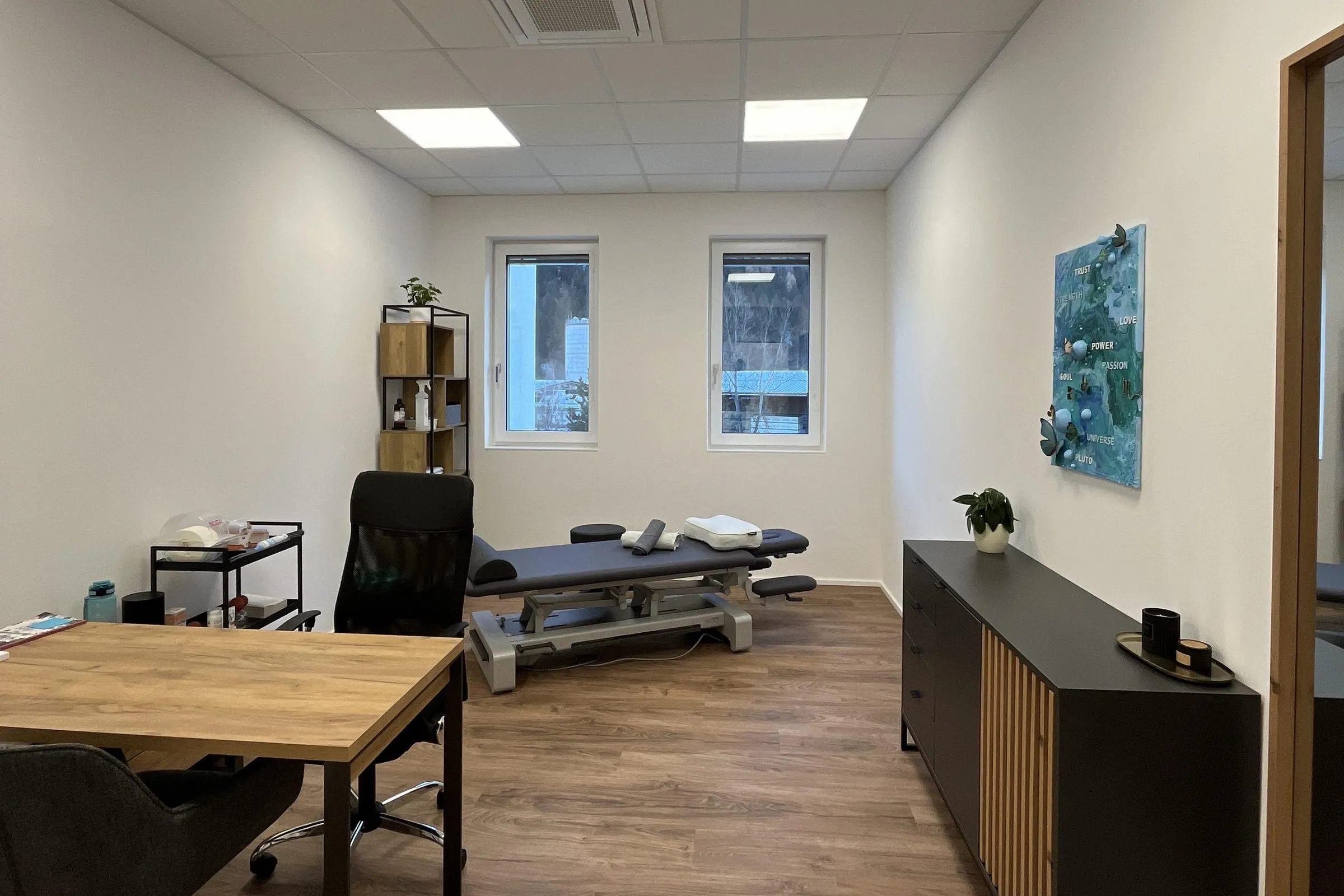 Behandlungsraum mit Therapieliege, Schreibtisch und zwei Fenstern