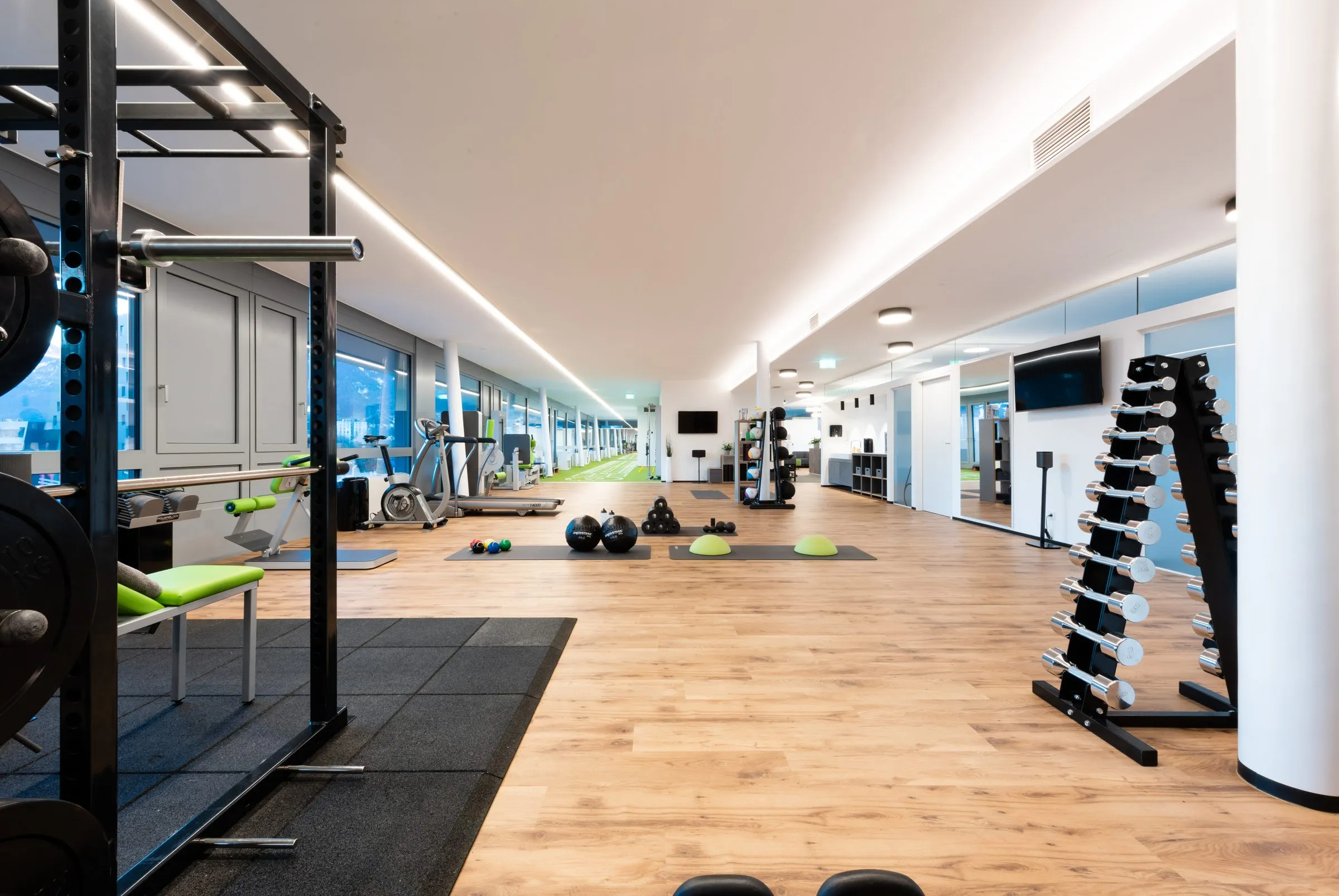 Heller Trainingsbereich mit Holzboden, Hantelständer, Ergometer und Trainingsgeräten entlang der Fensterfront