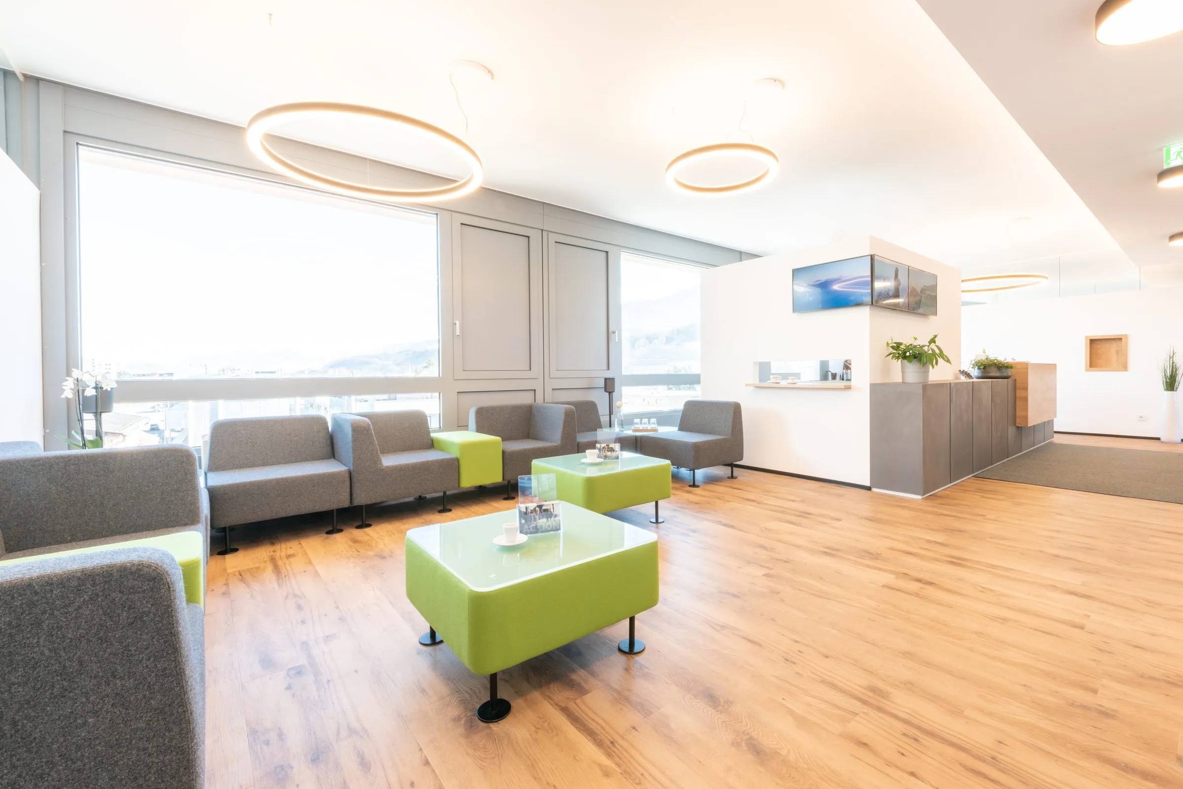 Wartebereich mit grauen Sofas, grünen Tischen und Empfang im Hintergrund